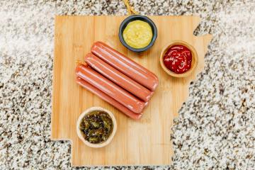 Raw natural beef and pork hot dogs on an Arkansas cutting board with ketchup, mustard, and relish flanking them